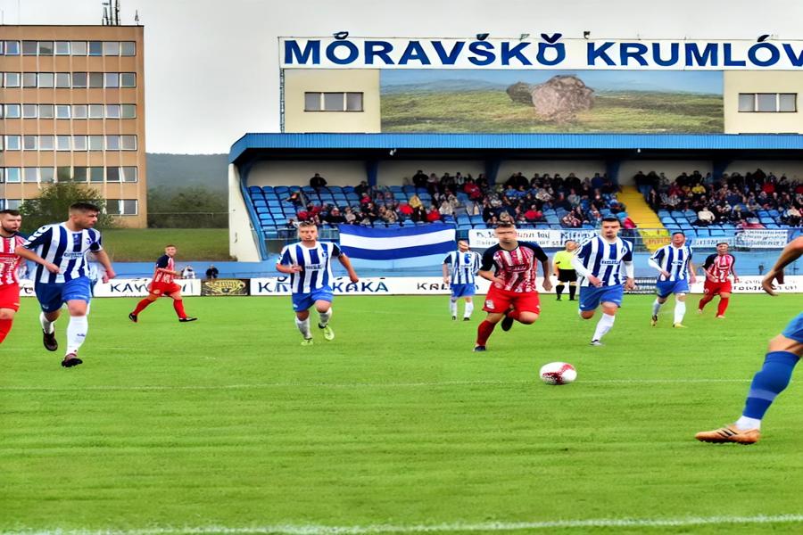 Fotbalový zápas ve sportovní aréně Moravského Krumlova. V pozadí je krumlovský sportovní areál, v modrobílých pruhovaných dresech Moravský Krumlov a v červených dresech tým Jaroměřic. Vytvořil Chat GPT